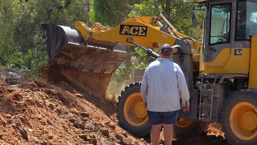 A man wearing a blue shirt standing next to an excavator pouring dirt