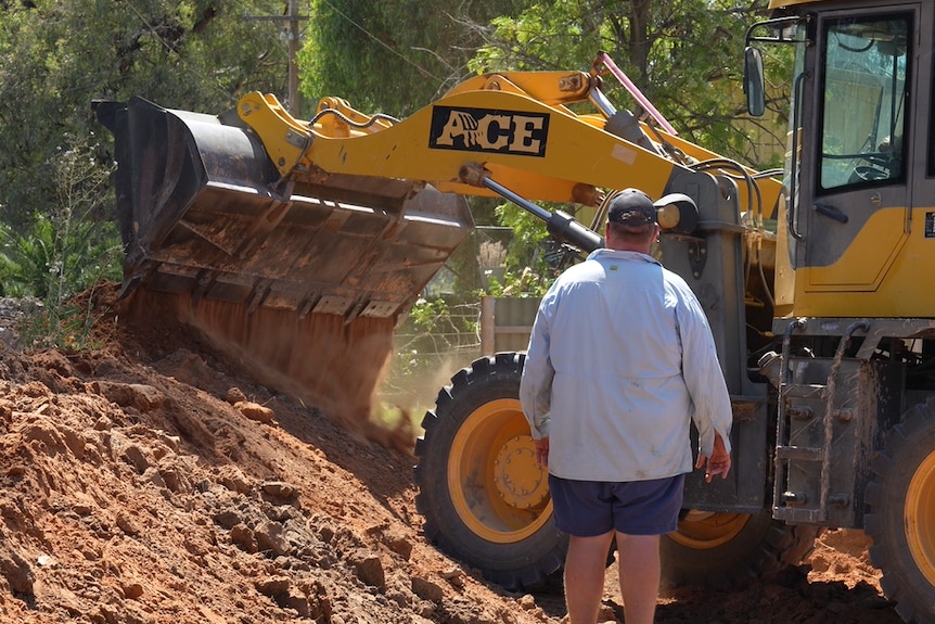 A man wearing a blue shirt standing next to an excavator pouring dirt