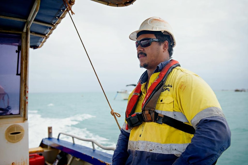 A man leans against a railing on a fishing-type boat, wearing a high-vis shirt, sunglasses and life jacket.