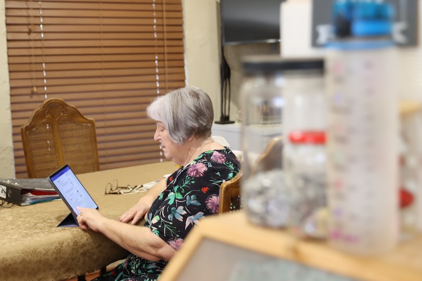 Older woman wearing floral top sits at a table looking at a tablet device.