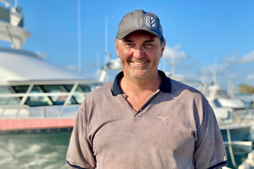 Un homme regarde la caméra, il porte un chapeau et il y a des bateaux en arrière-plan.