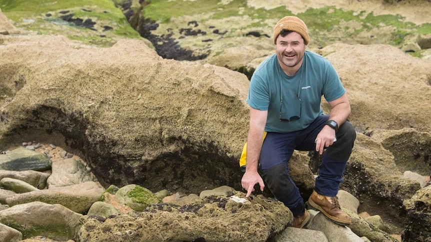 A man stand among several rocks, pointing at a possible megafaunal kangaro fossil in a rock