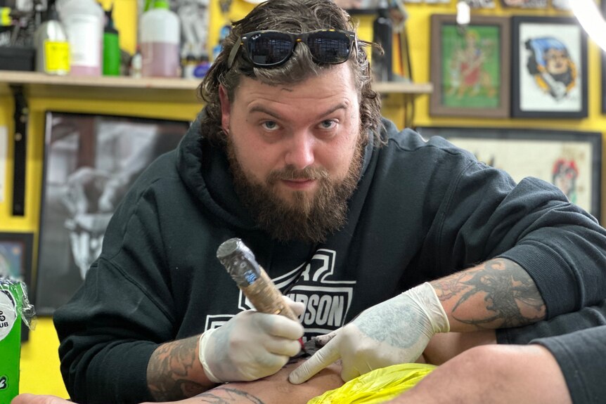 A man gives a tattoo on another man's leg while looking at the camera.