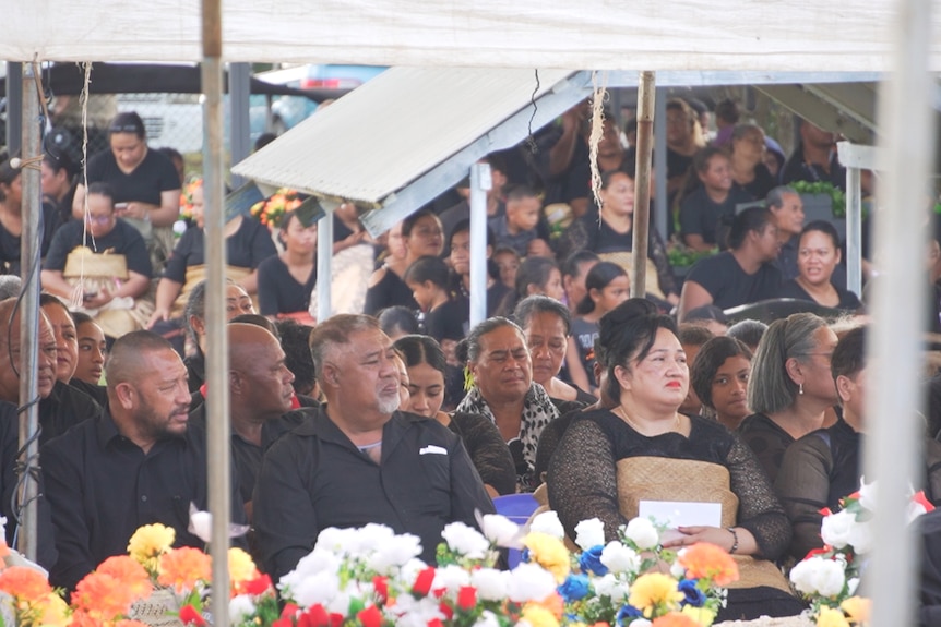 A crowd view of the funeral attendees. Flowers are shown, and many wearing black.