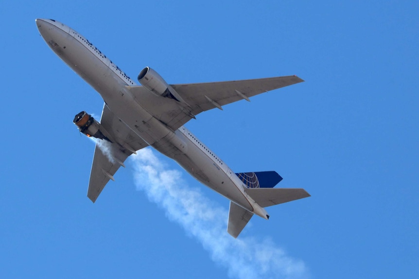 A silver passenger jet blows smoke from its left-hand engine as it flies beneath a blue sky.