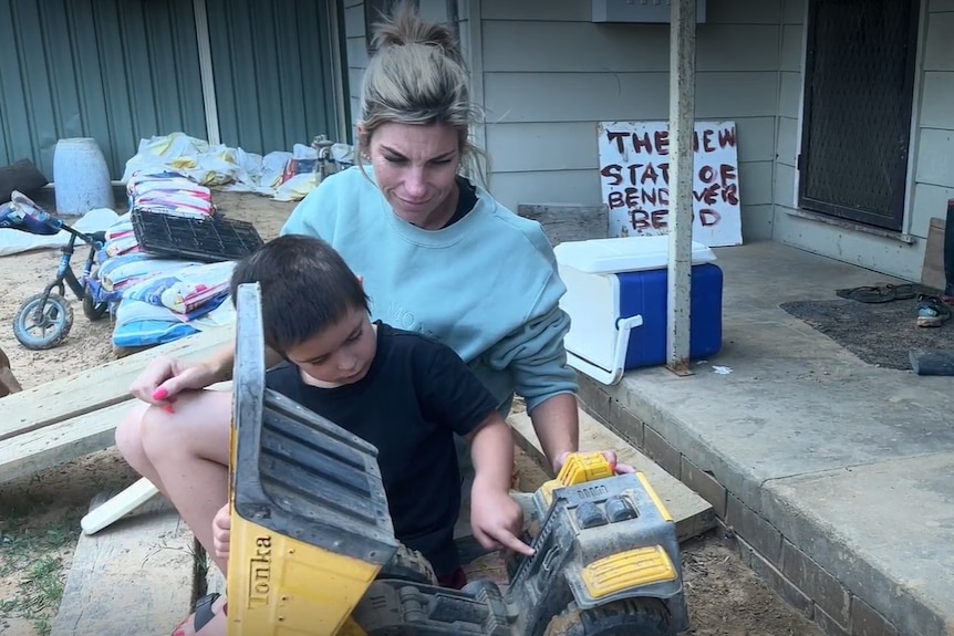 A woman and a boy sit on a step at a house that has been damaged by flooding.