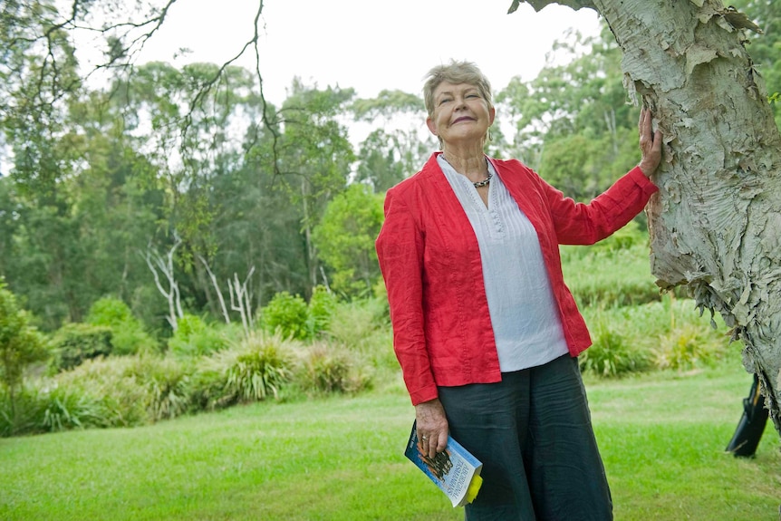 Historian Lyndall Ryan leans against a tree holding a book.