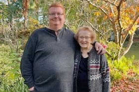 A middle-aged, ginger-haired man stands in a garden with his arm around an elderly woman.