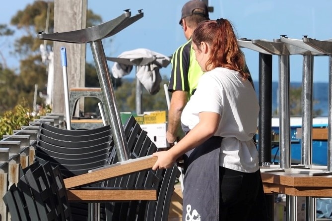 A young woman wearing an apron stacks a cafe table on top of another.