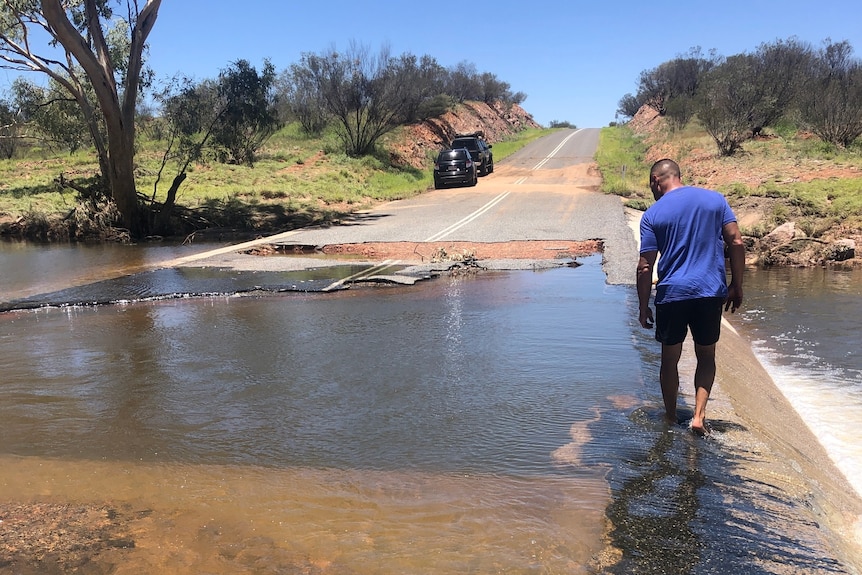 Water runs across badly damaged road with man in blue t-shirt standing on it and two cars parked in distance