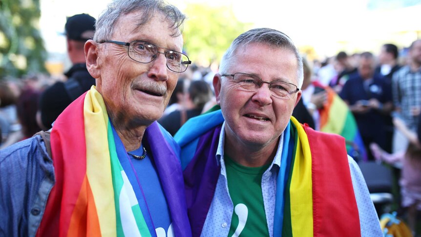 Geoff Bishop and Neil Connery stand side by side smiling with rainbow flags around their neck.
