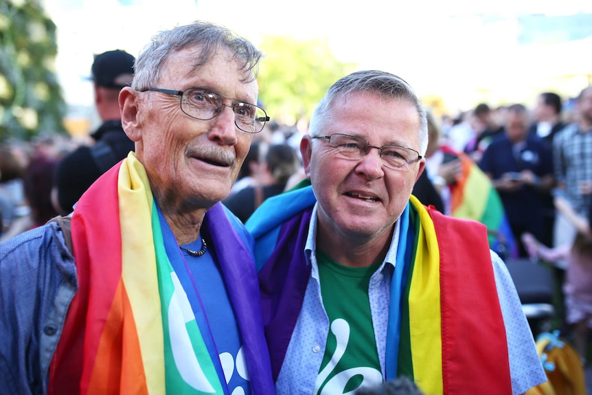 Geoff Bishop and Neil Connery stand side by side smiling with rainbow flags around their neck.