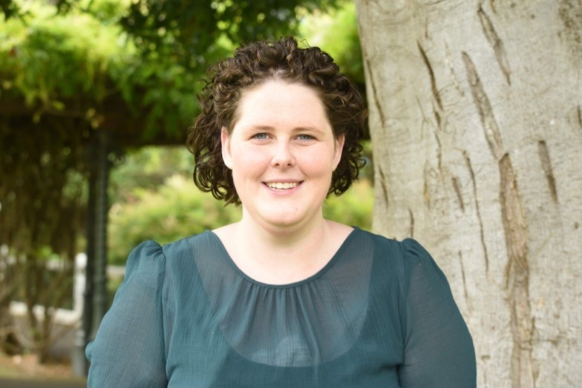 Julia wears a green shirt and smiles in front of a tree.