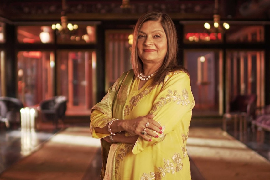 A still of a woman with long brown hair and yellow dress smiling to camera.