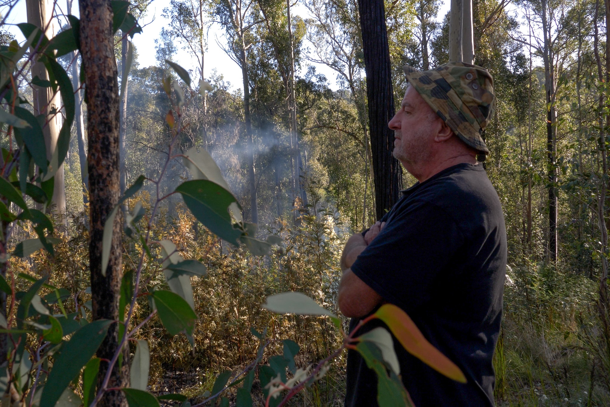 Profile of David with hat standing in bush land with smoke in the near distance.