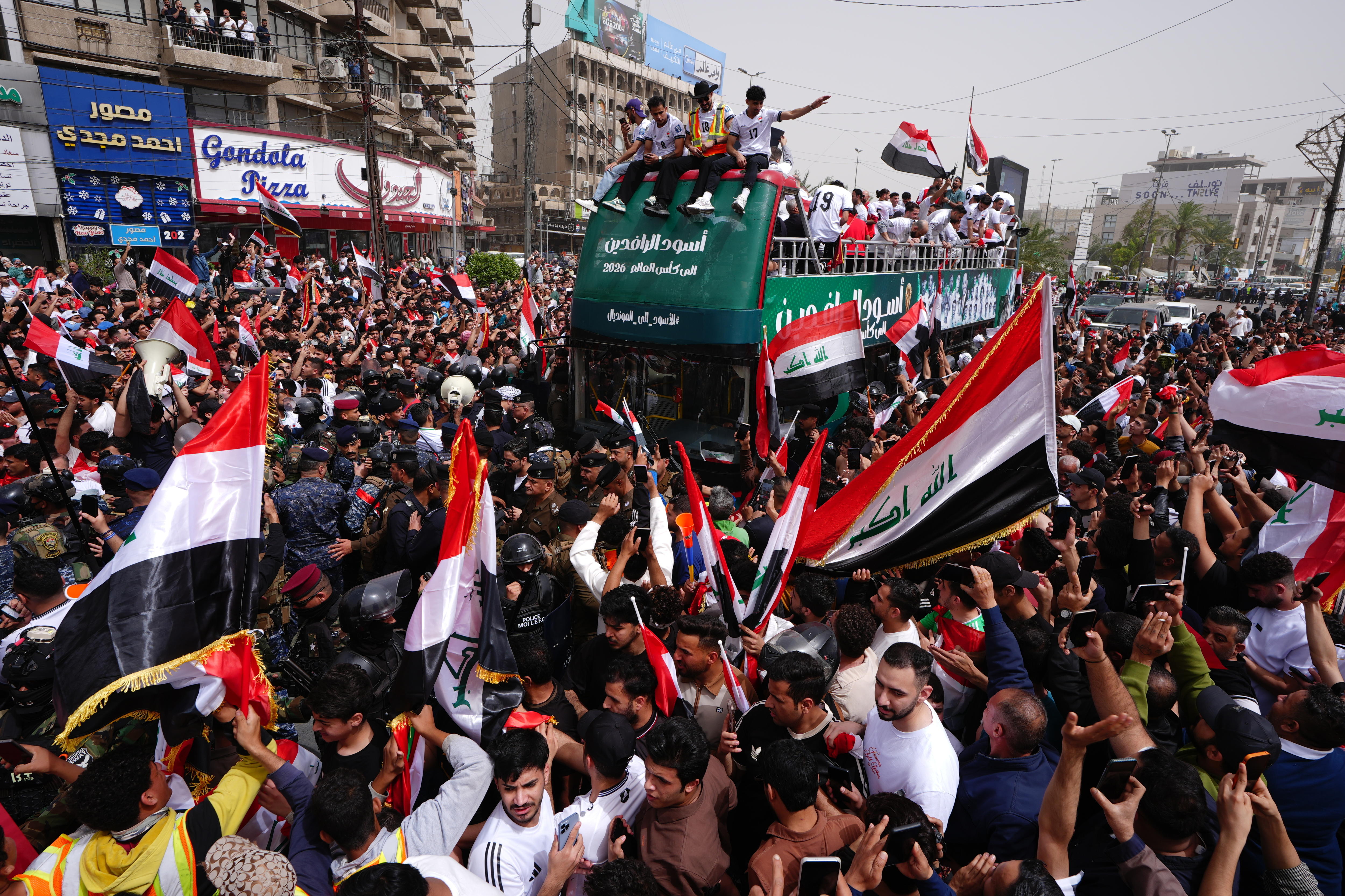 Members of Iraq's men's football team are on an open top bus, surrounded by fans waving Iraqi flags