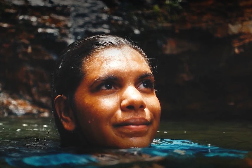 A young Indigenous girl swims in a water hole smiling.