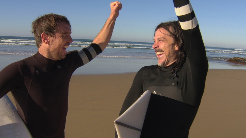 Two men in wetsuits fist pump the air with glee at the beach