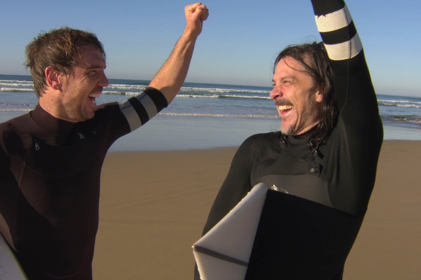 Two men in wetsuits fist pump the air with glee at the beach