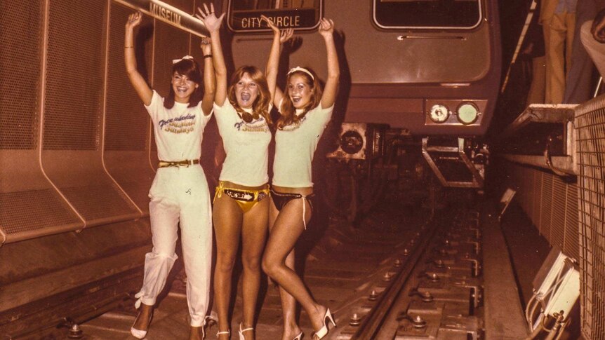 Three women in bikinis and high heels stand in a rail tunnel in front of a train