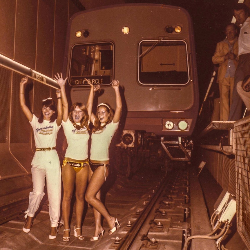 Three women in bikinis and high heels stand in a rail tunnel in front of a train