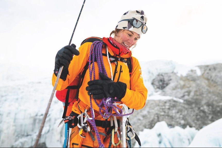 A woman in bright orange mountain-climbing gear smiles on top of a snow peak.