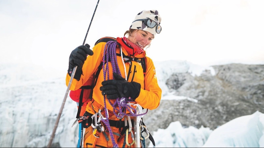 A woman in bright orange mountain-climbing gear smiles on top of a snow peak.