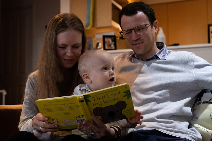 A baby boy holds a book while sitting in the laps of his parents on a couch.