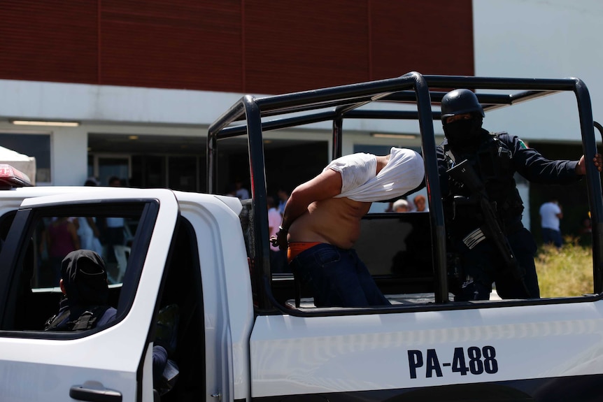 Police escort a man with a white shirt covering his head into a police vehicle.