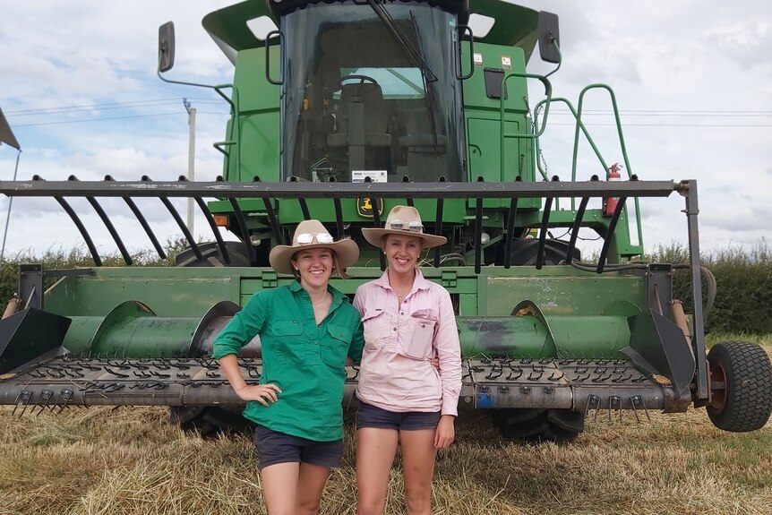 two young women stand in front of a grain harvester