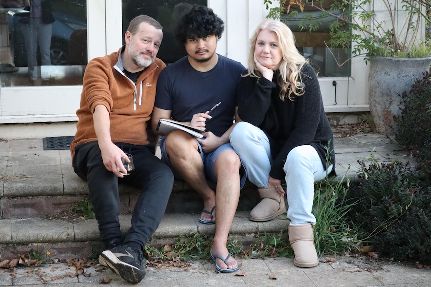 A man and his wife sit on a step of their home with their adult son who sits with a drawing pad and pen