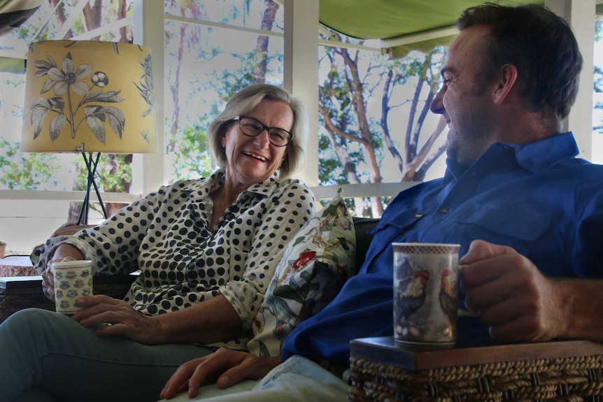 Ann and Rupert Ballinger look at each other, sitting side by side on a couch with mugs of tea.
