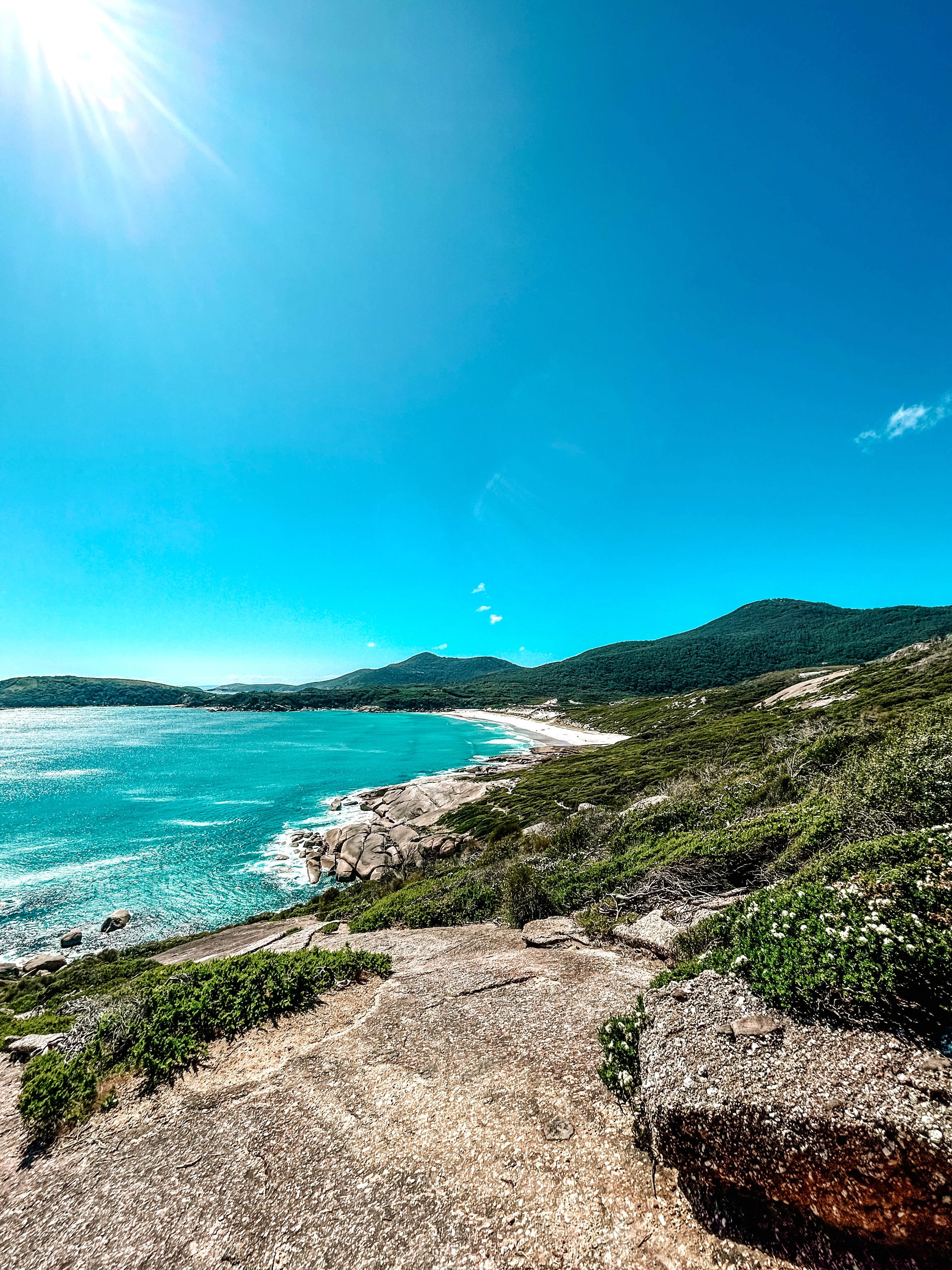 Bare boulders surrounded by green vegetation slope down to the aqua coloured water of the beach with green mountains behind.
