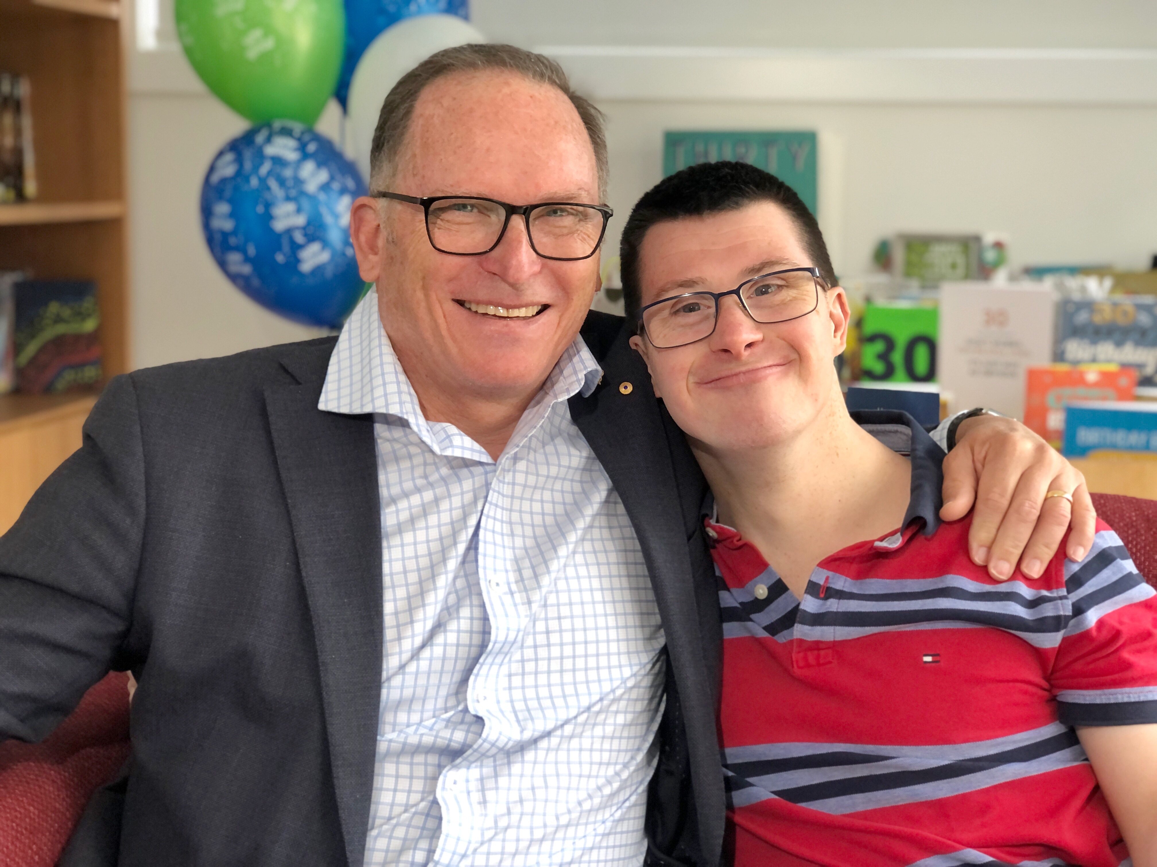 A man with with grey hair smiles with his arm around a younger man with dark hair who is also smiling.