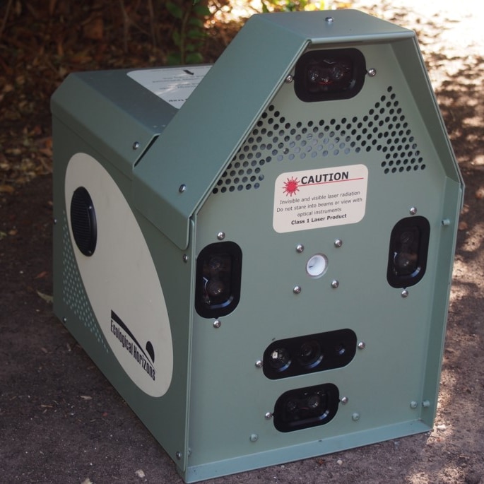 A green machine with sensors on the front sits on the ground surrounded by leaves and dirt.