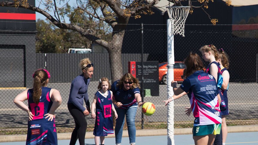 A group of netball players stand around a goal as a little girl -flanked by two adults- throws the ball from the sidelines