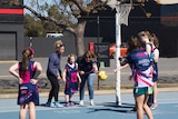 A group of netball players stand around a goal as a little girl -flanked by two adults- throws the ball from the sidelines