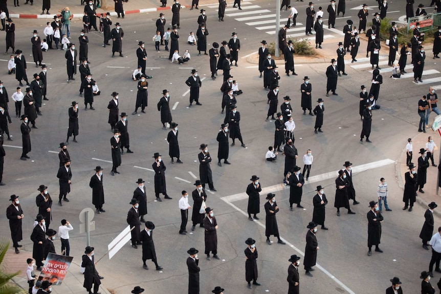 A number of menand children  dressed in black suits and wearing masks with hats on stand in a street.
