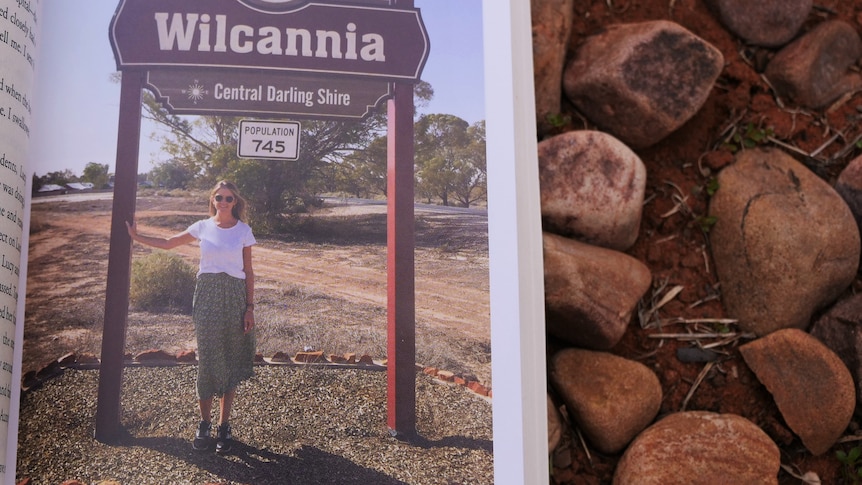 A photo of a page of a book which includes Sarah Donnelley in front of the Wilcannia sign against a bed of rocks