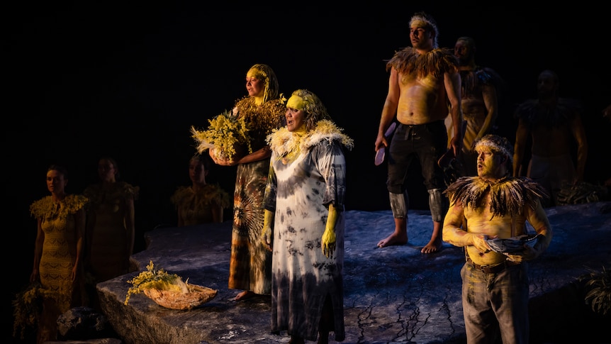 A group of Indigenous performers stand on stage wearing yellow ochre and holding wattle, singing.