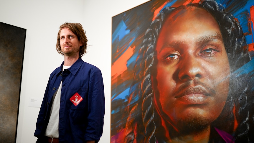 Matt Adnate stands in front of a photorealistic portrait of Baker Boy, a young Aboriginal man.