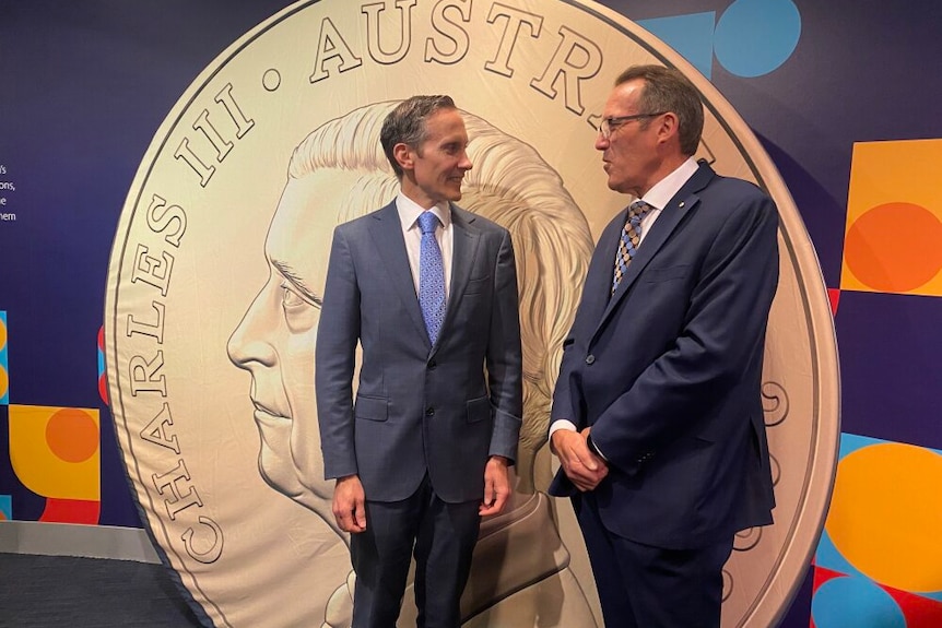 Two men in front of an oversized coin with the king's face on it