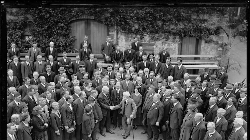 A black and white photo of a large group of people in a garden, mainly men in suits, surrounding two men shaking hands.