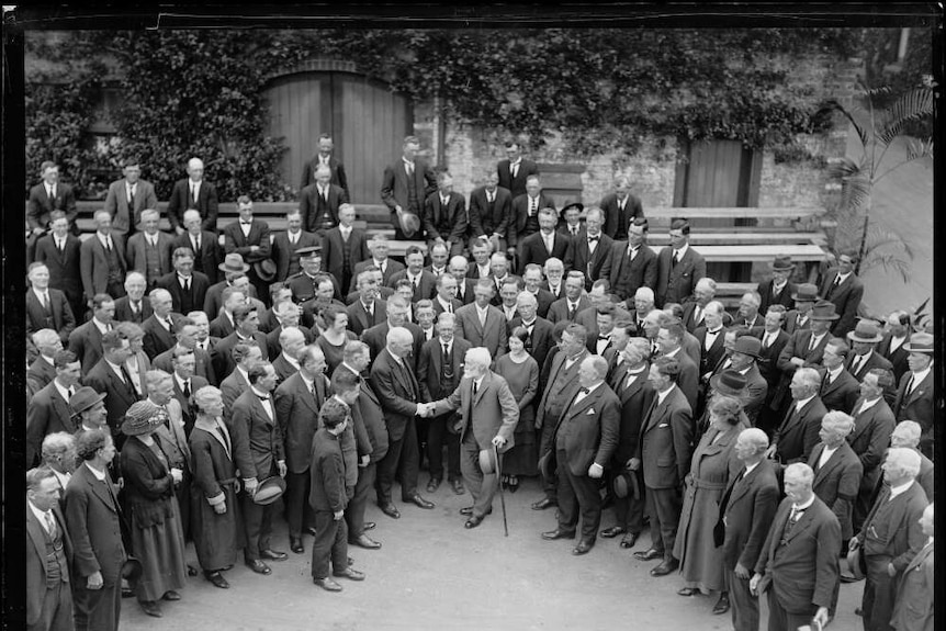 A black and white photo of a large group of people in a garden, mainly men in suits, surrounding two men shaking hands.