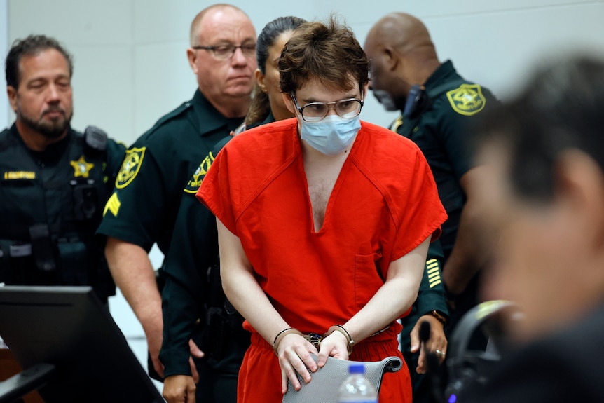 A handcuffed prisoner in an orange jumpsuit and face mask is escorted into a courtroom by officials.