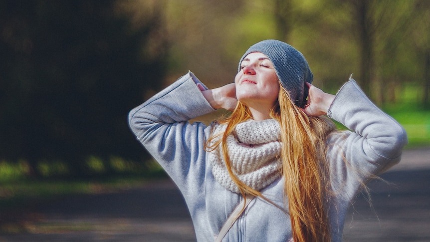 A woman wearing a beanie, a chunky scarf and a grey winter jacket is standing in a park enjoying the winter sun.