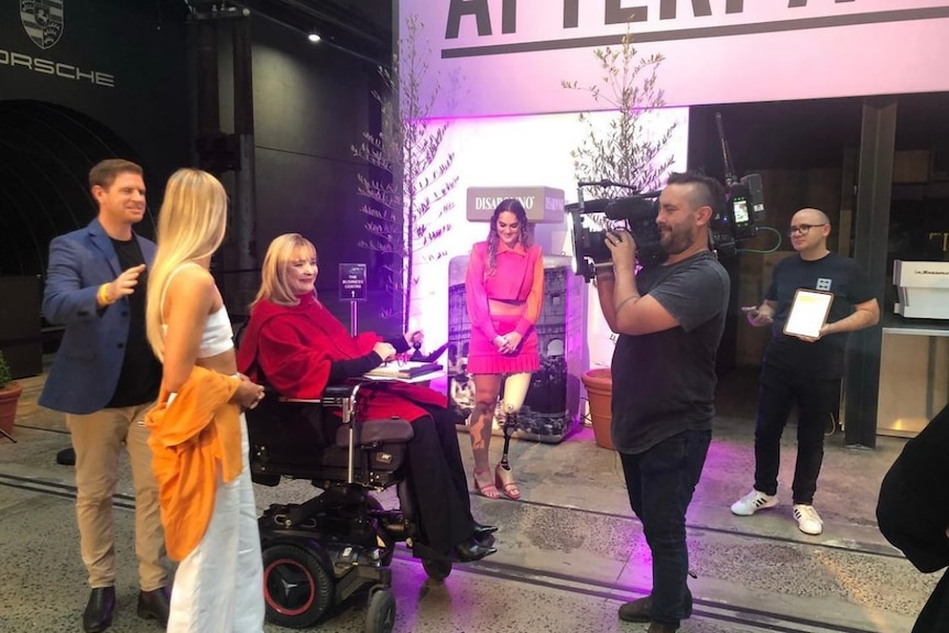 A woman in a wheelchair standing next to two women, being filmed by a camera operator
