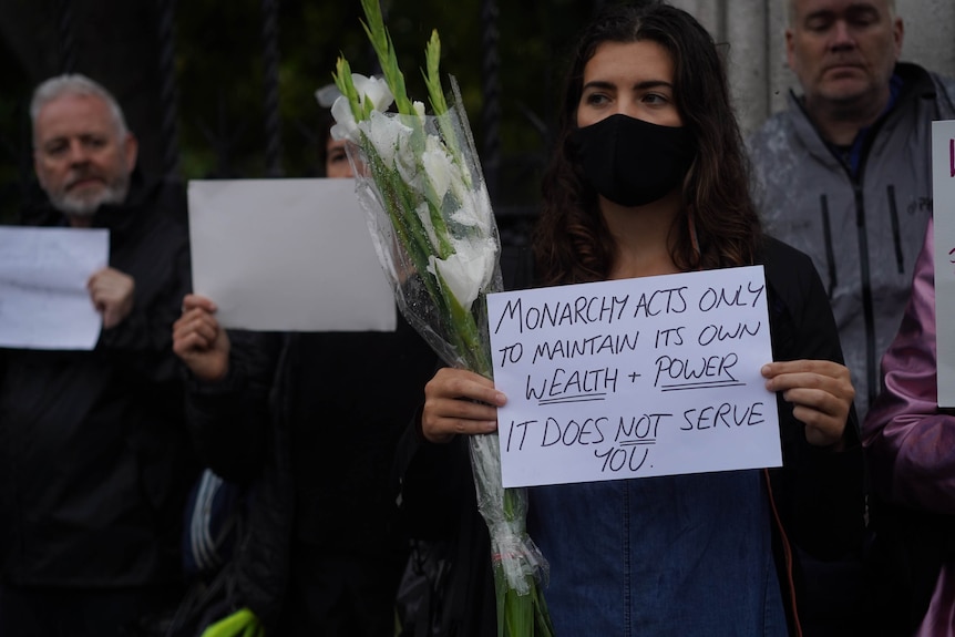 A woman holds a sign that says 'monarchy acts to maintain its wealth' 