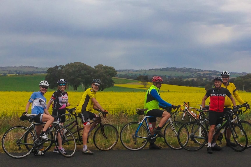 A group of cyclists with bikes and wearing lycra, standing in front of a paddock of yellow canola