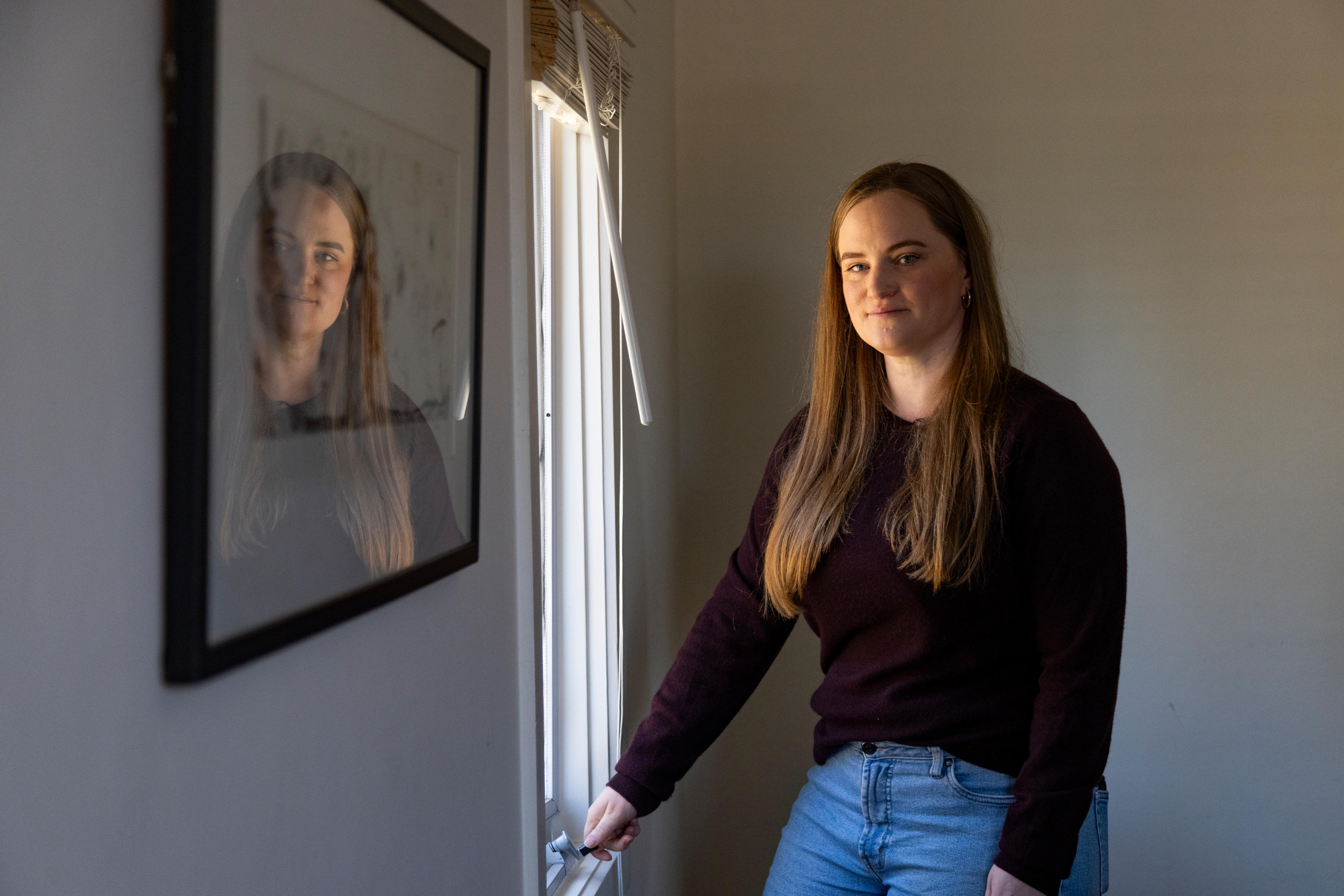 A woman wearing black stands by a window, her reflection visible in a picture frame.